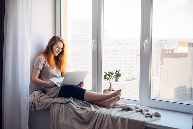 Belle fille positive utilisant un ordinateur portable, assise sur le rebord de la fenêtre dans un appartement en ville. Jeune femme rousse travaillant à la maison. Notion d'indépendant.