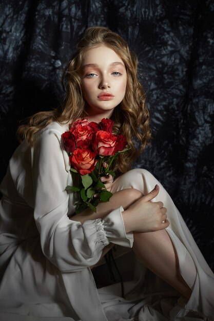 Belle fille de printemps est assise sur le sol avec des fleurs roses dans ses mains. Une femme en robe blanche rêve, une image romantique. Blonde aux cheveux bouclés