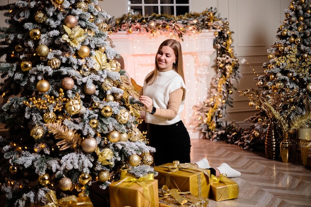 Une belle fille en pull est assise près d'un arbre de Noël décoré de guirlandes À côté se trouve un arbre de Noël avec des boîtes de cadeaux décorés du Nouvel An