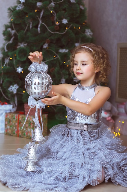 Une belle fille en robe élégante est assise près du sapin de Noël et tient une décoration de Noël