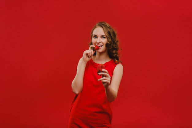 Une belle fille en robe rouge sur fond rouge tient une fraise dans ses mains et sourit