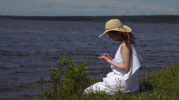 Belle fille rousse dans une robe d'été blanche utilise un téléphone portable assis sur le rivage et les rivières