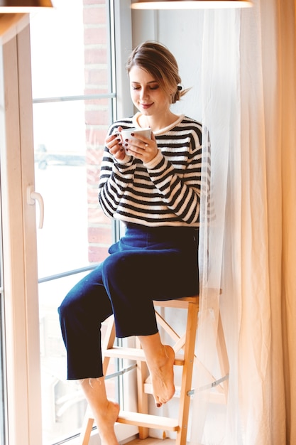 Belle fille rousse avec une tasse de café est assise sur de petits escaliers à la maison près de la fenêtre.