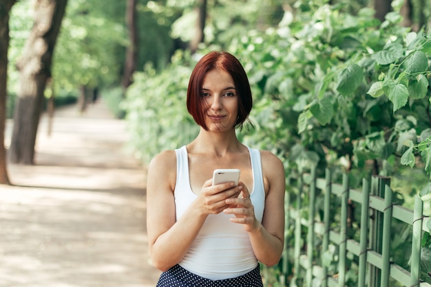 Belle fille rousse tenant un smartphone et regardant la caméra dans le parc en été.
