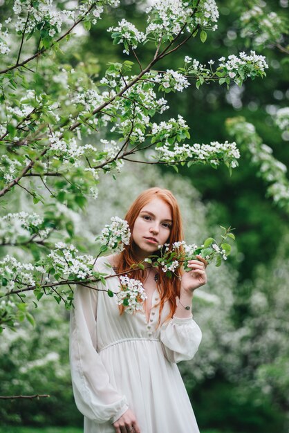 Belle fille rousse vêtue d'une robe blanche au milieu des pommiers en fleurs dans le jardin.