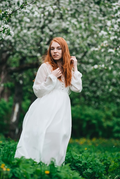 Belle fille rousse vêtue d'une robe blanche au milieu des pommiers en fleurs dans le jardin