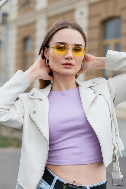 Belle fille de rue hipster à la mode avec des lunettes de soleil jaunes élégantes et un t-shirt violet avec une veste en cuir dans la ville