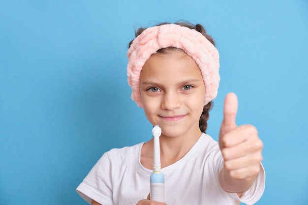 Belle fille se brosse les dents avec une brosse à dents électrique et fait des pouces vers le haut sur un gros plan de fond bleu. Le concept d'hygiène et de soins bucco-dentaires chez un enfant.