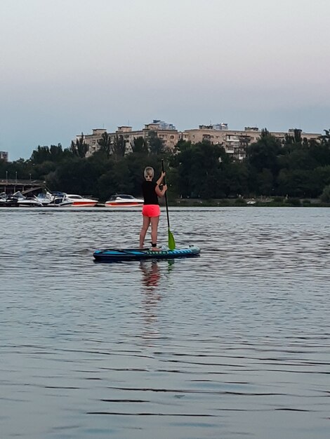 Une belle fille se tient sur une planche de natation et tient une rame au-dessus de sa tête dans le contexte de la rivière et de la forêt et du soleil éclatant