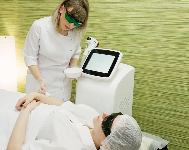 Une belle fille se trouve dans le bureau d'une esthéticienne dans des lunettes d'irradiation laser les mains du médecin enlèvent les cheveux sur les mains avec un appareil laser