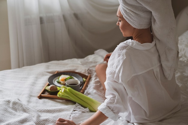 Belle fille avec une serviette blanche sur la tête est assise dans le lit et va prendre son petit déjeuner