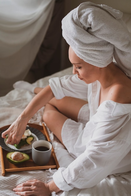 Belle fille avec une serviette blanche sur la tête est assise dans son lit et prend son petit déjeuner avec un sandwich sain avec du café