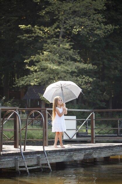 Une belle fille de six ans vêtue d'une courte robe blanche se dresse sur la maçonnerie près de la rivière et tient un parapluie blanc dans ses mains