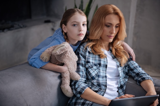 Belle fille solitaire bouleversée attendant l'attention de la mère à la maison et étreignant le parent pendant que la mère surfe sur Internet et utilise la tablette