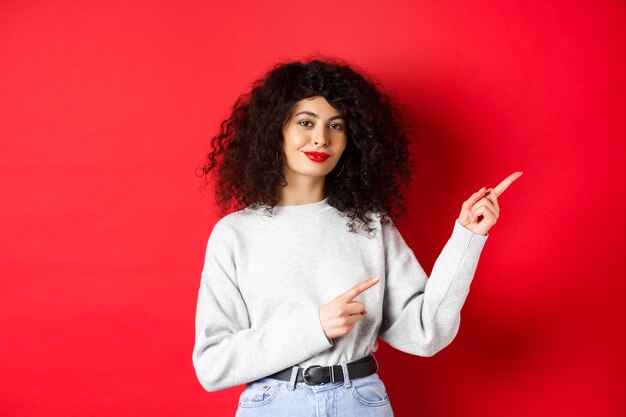 Belle fille souriante aux cheveux bouclés, pointant du doigt le logo, montrant une publicité, fond rouge