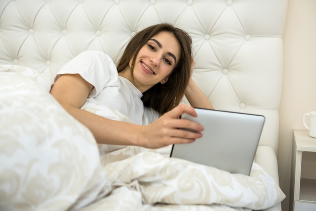 Belle fille souriante couchée dans son lit le matin se détendre sur sa journée de jeu sur une tablette