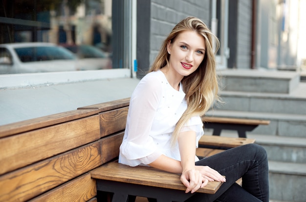 Belle fille souriante heureuse avec de longs cheveux blonds, des lèvres rouges, une chemise blanche qui pose dans la rue un jour d'été.