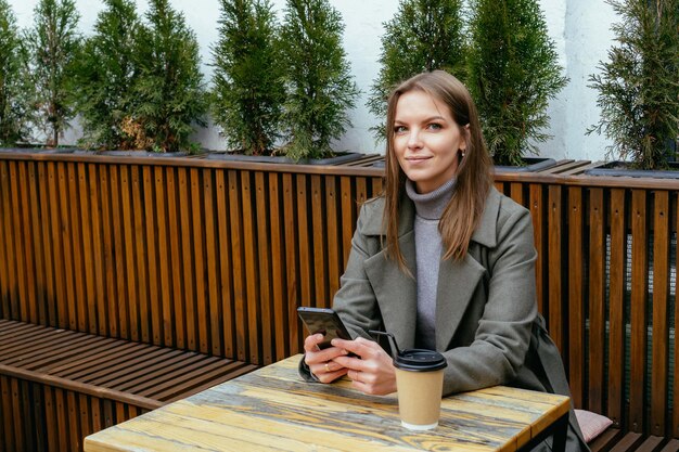 Belle fille souriante avec des lunettes assise dans un café tenant un smartphone dans les mains et discutant
