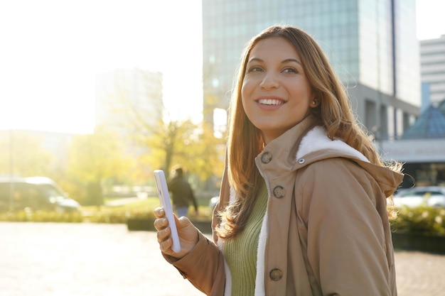 Belle fille souriante tenant un smartphone à l'extérieur au coucher du soleil