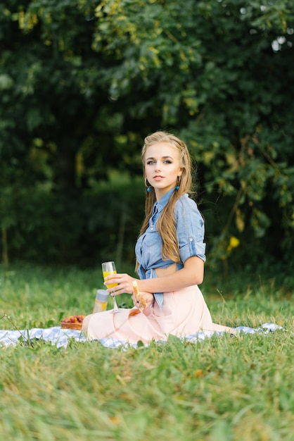 Une belle fille souriante tient un petit pain frais dans ses mains et s'assoit sur une couverture en plein air dans le parc. Pique-nique d'été dans le parc