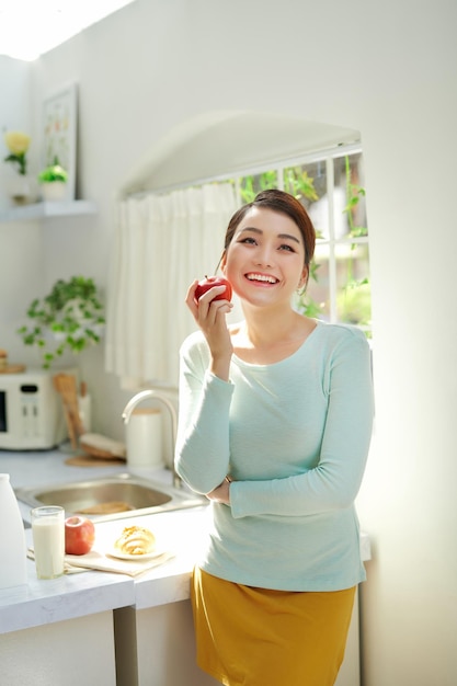 Belle fille tenant une pomme rouge en se tenant debout dans la cuisine de la maison le matin