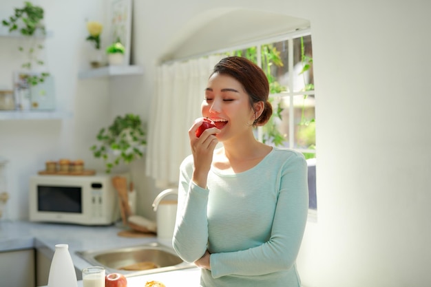 Belle fille tenant une pomme rouge en se tenant debout dans la cuisine de la maison le matin