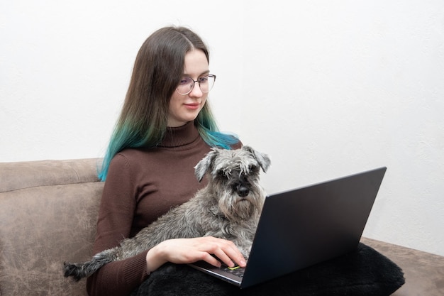 Une belle fille en tenue décontractée utilise un ordinateur portable et sourit tout en étant assise avec son chien mignon sur le canapé en gros plan L'animal regarde la caméra Étudiante étudiant en ligne avec son chien