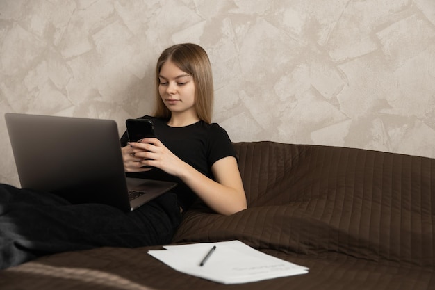 Belle fille travaille à la maison à l'ordinateur allongé sur le lit et vérifie les messages au téléphone