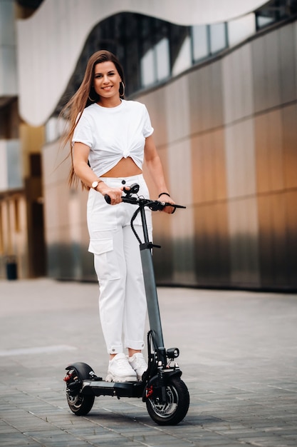 Belle fille en vêtements élégants blancs sur un scooter électrique dans la ville.