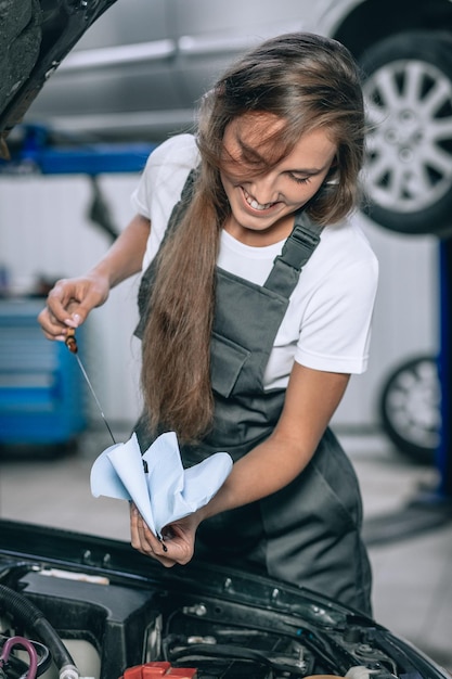 Une belle fille vêtue d'une combinaison noire et d'un t-shirt blanc sourit, vérifiant le niveau d'huile dans une voiture noire dans le garage.