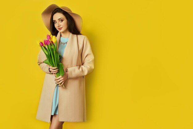 Belle fille vêtue d'un manteau et d'un chapeau avec des tulipes dans les mains sur fond jaune