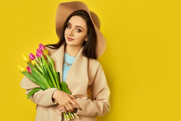 Belle fille vêtue d'un manteau et d'un chapeau avec des tulipes dans les mains sur fond jaune