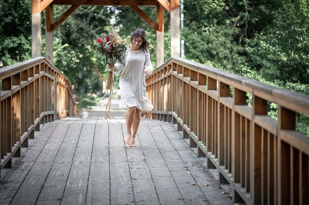 Belle fille vêtue d'une robe blanche avec un bouquet de fleurs exotiques sur un pont en bois.
