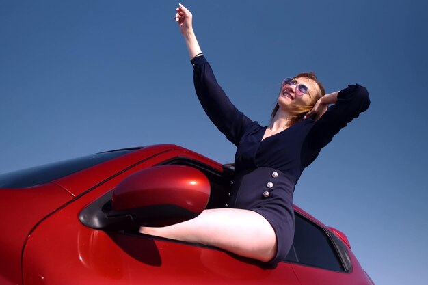 Photo belle fille vêtue d'une robe courte noire et de lunettes de soleil est assise penchée par la fenêtre d'une voiture rouge s'amusant et dansant