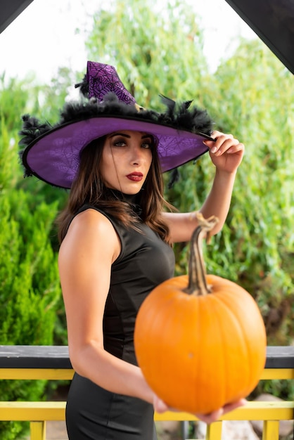 Belle fille vêtue d'une robe noire et d'un chapeau de sorcière pose dans un belvédère Tient une citrouille dans ses mains Halloween