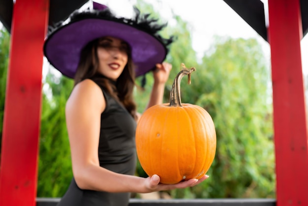 Belle fille vêtue d'une robe noire et d'un chapeau de sorcière pose dans un belvédère Tient une citrouille dans ses mains Halloween