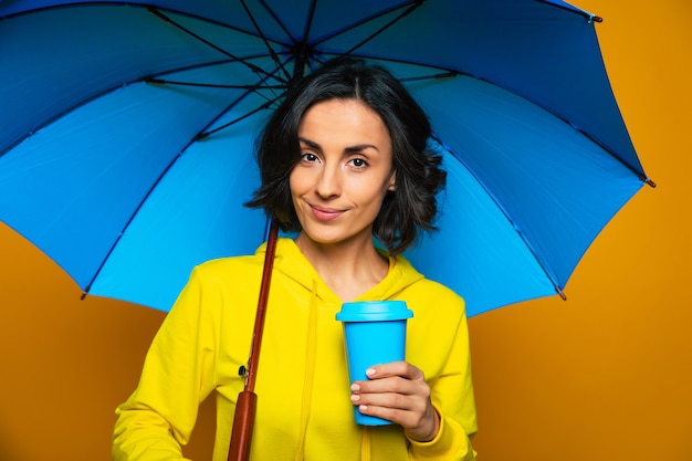 belle fille vêtue d'un sweat à capuche jaune sous un parapluie, tenant une tasse bleue dans sa main gauche, s'amusant.
