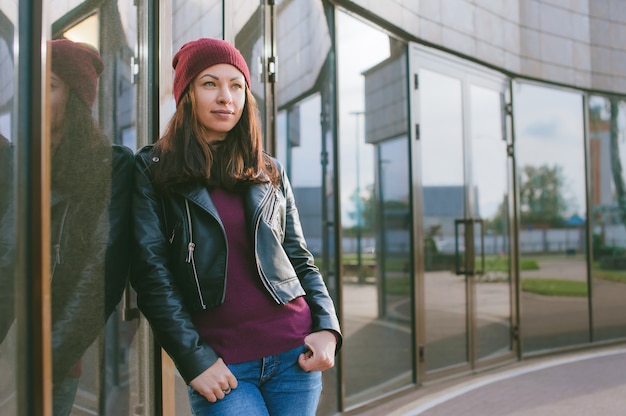 Une belle fille vêtue d'une veste en cuir et d'un chapeau se dresse sur fond de bâtiment en verre.