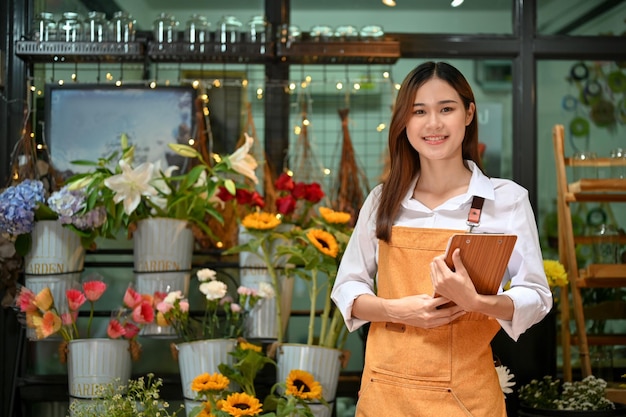 Belle fleuriste asiatique se tient devant l'entrée de son magasin de fleurs tenant un presse-papiers