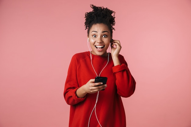 belle incroyable jeune femme africaine excitée et heureuse posant isolée sur un mur rose écoutant de la musique avec des écouteurs à l'aide d'un téléphone portable.