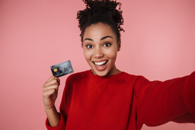 belle incroyable jeune femme africaine excitée heureuse posant isolée sur mur rose prendre selfie par caméra avec carte de crédit.