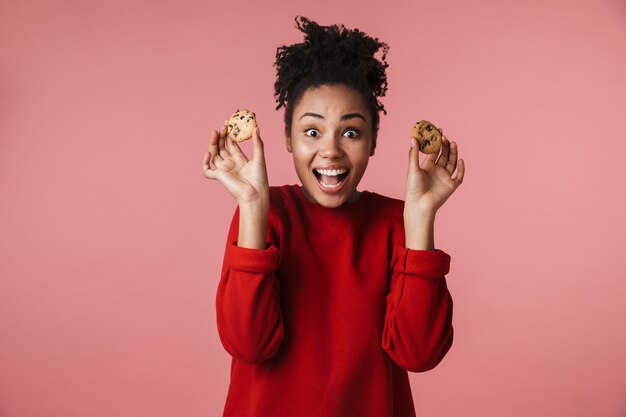 belle incroyable jeune femme africaine excitée heureuse posant isolée sur un mur rose tenant des biscuits sucrés.