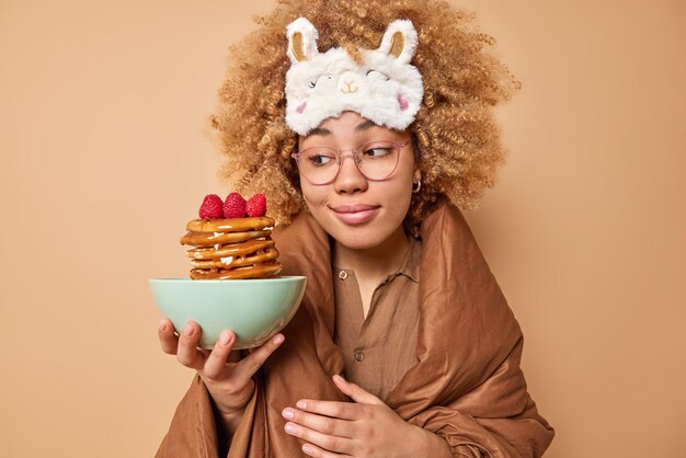 Belle jeune belle femme aux cheveux bouclés et touffus porte un masque de sommeil regarde des crêpes appétissantes avec du sirop et des framboises enveloppées dans une couette a un délicieux petit déjeuner isolé sur fond beige