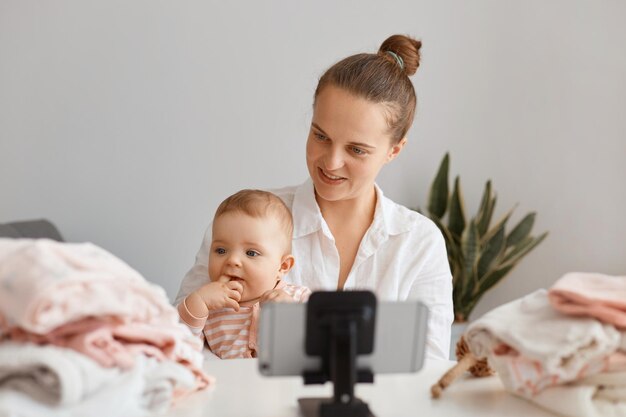 Belle jeune blogueuse adulte assise à table avec un enfant en bas âge et enregistrant une vidéo pour son vlog, utilisant un téléphone portable sur un trépied, créant du contenu avec sa fille.