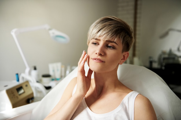 Belle jeune blonde aux cheveux courts touchant doucement son visage alors qu'elle était assise sur une chaise dans un salon de beauté dans une clinique spa. Concept de soins de la peau