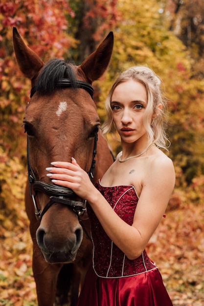 Belle jeune blonde vêtue d'une robe bordeaux embrasse un cheval bai