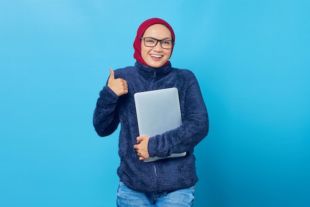 Belle jeune étudiante joyeuse portant une veste bleue tenant un ordinateur portable et donnant des pouces vers le haut