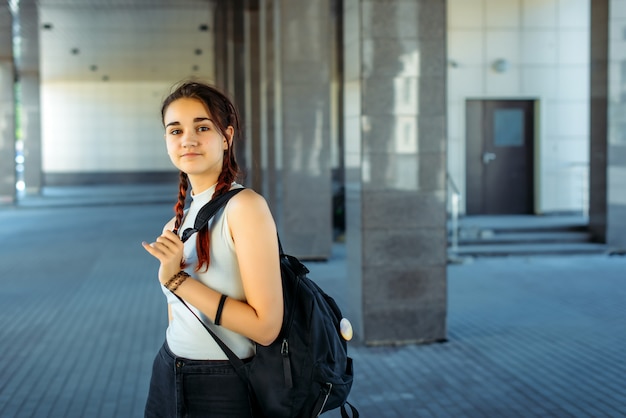 Belle jeune étudiante avec sac à dos sur l'épaule va à l'école, gros plan. Écolière avec deux tresses debout devant le collège