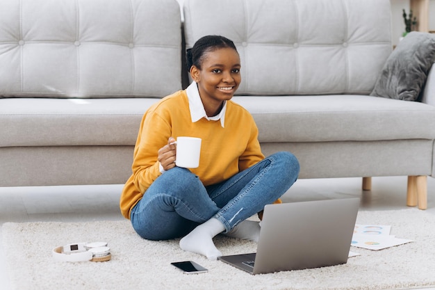 Belle jeune étudiante universitaire noire assise sur le sol, buvant du café et faisant un projet sur un ordinateur portable