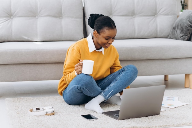 Belle jeune étudiante universitaire noire assise sur le sol en buvant du café et en faisant un projet sur un ordinateur portable
